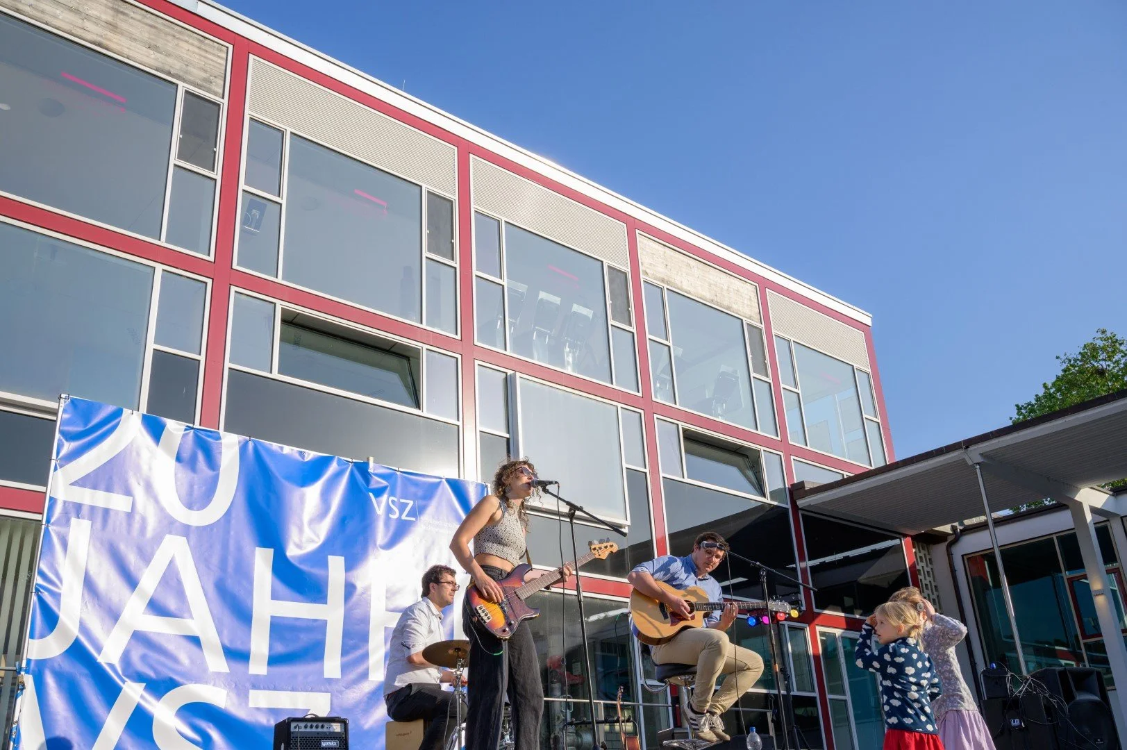 Musiker spielen auf einer Bühne im Freien vor einem modernen Gebäude mit großem Glasfenster. Es gibt Kinder, die der Performance zuschauen.
