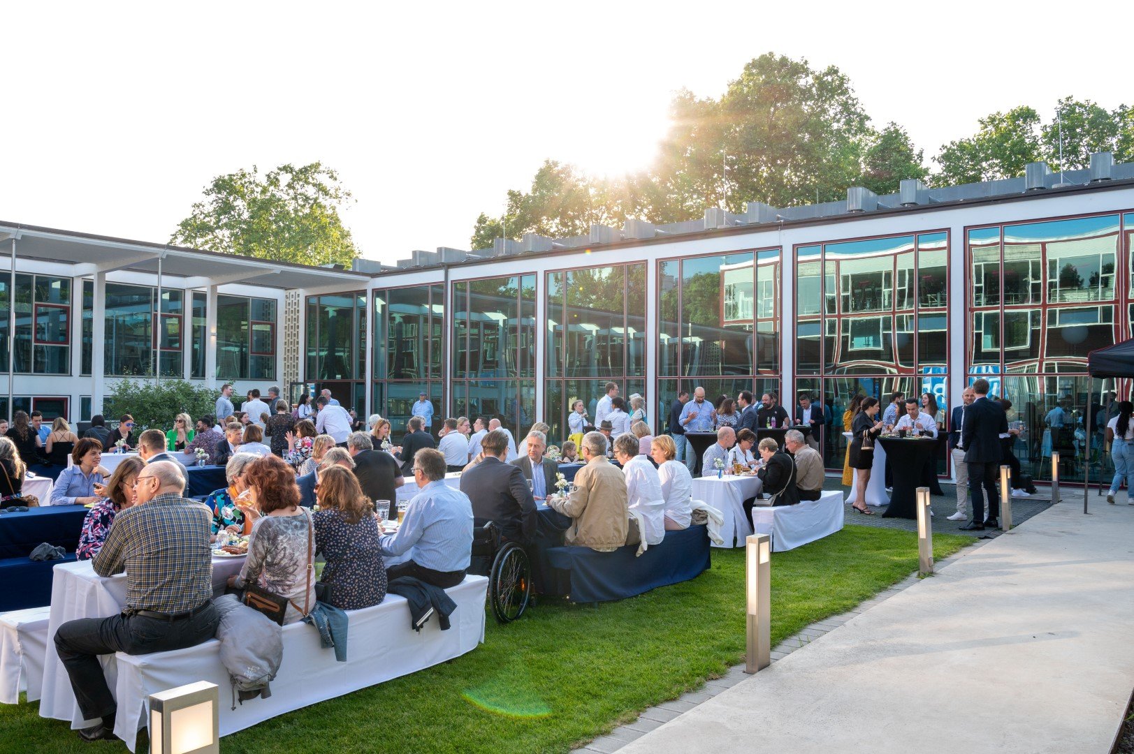 Außenveranstaltung mit vielen Menschen, Tischen und einem modernen Glasgebäude im Hintergrund, Sonnenlicht und grüne Bäume.