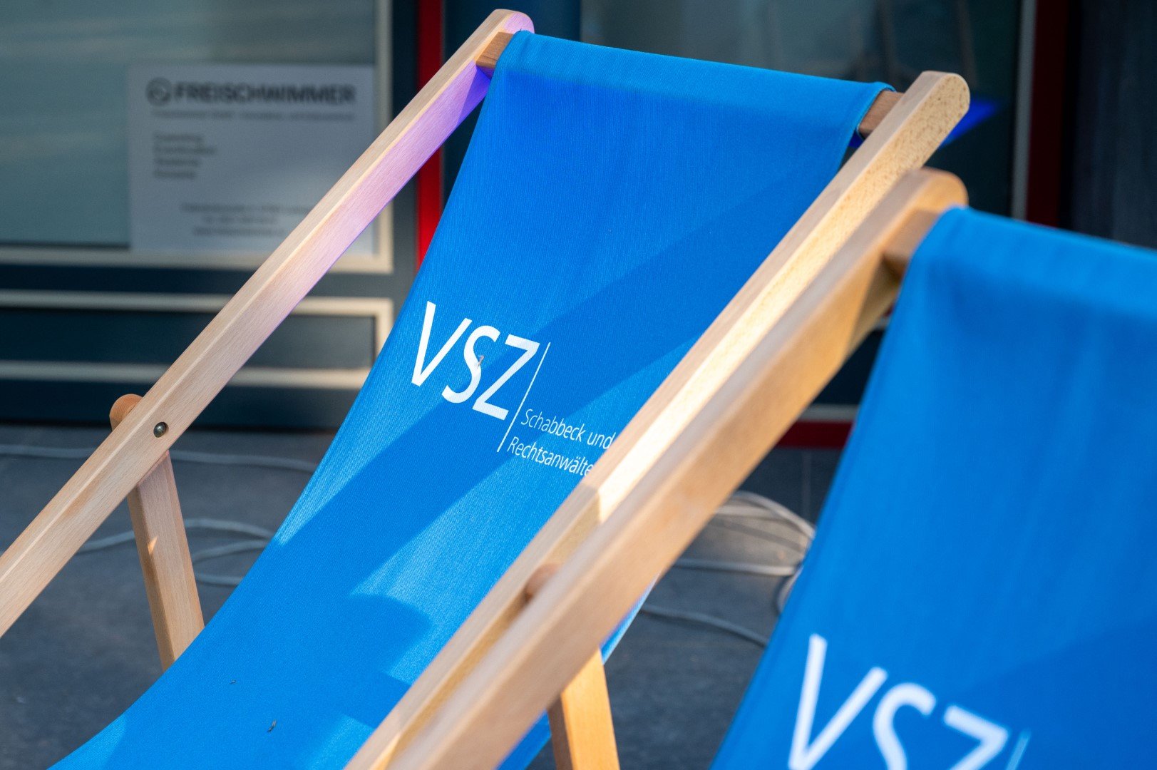 Zwei Sonnenliegen mit blauen Stoffbezügen, die den Schriftzug 'VSZ' und weitere kleinere Texte tragen, vor einem Gebäude.