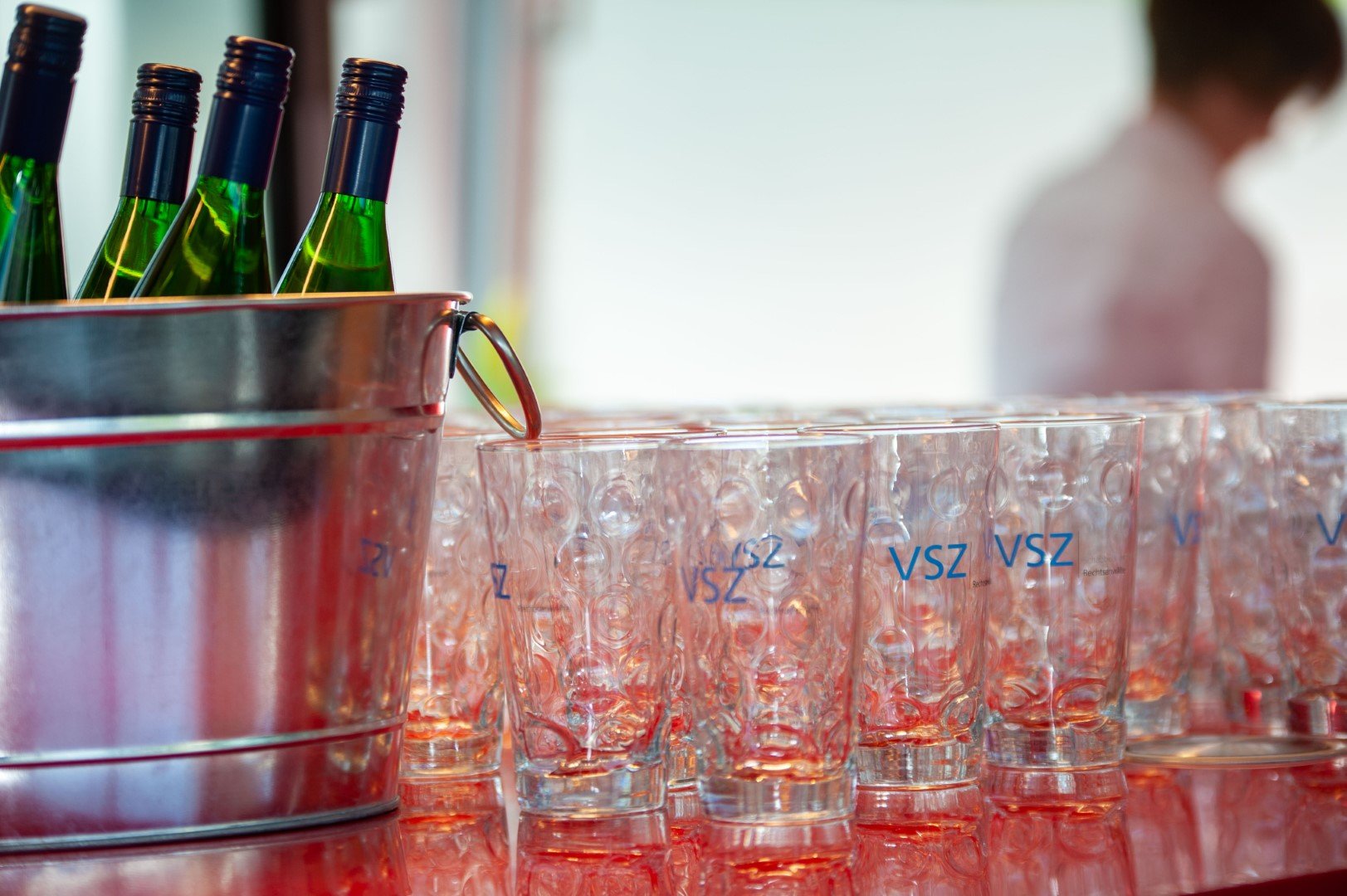 Mehrere leere Gläser mit blauer Aufschrift 'VSZ' nebeneinander auf einem Tisch, daneben ein Metallbehälter mit grünem Wein in Flaschen.