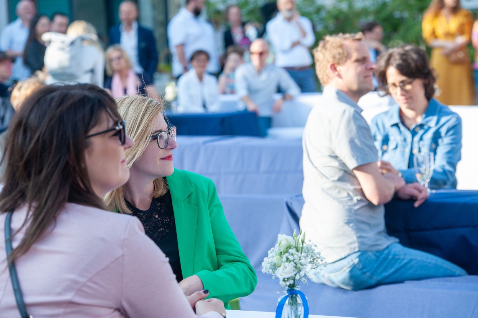 Mehrere Menschen sitzen und stehen bei einer Outdoor-Veranstaltung. Im Vordergrund sind zwei Frauen mit Brille zu sehen, eine trägt eine grüne Jacke. Im Hintergrund sprechen andere Menschen, einige sitzen an Tischen, und es gibt einen Tisch mit Blume