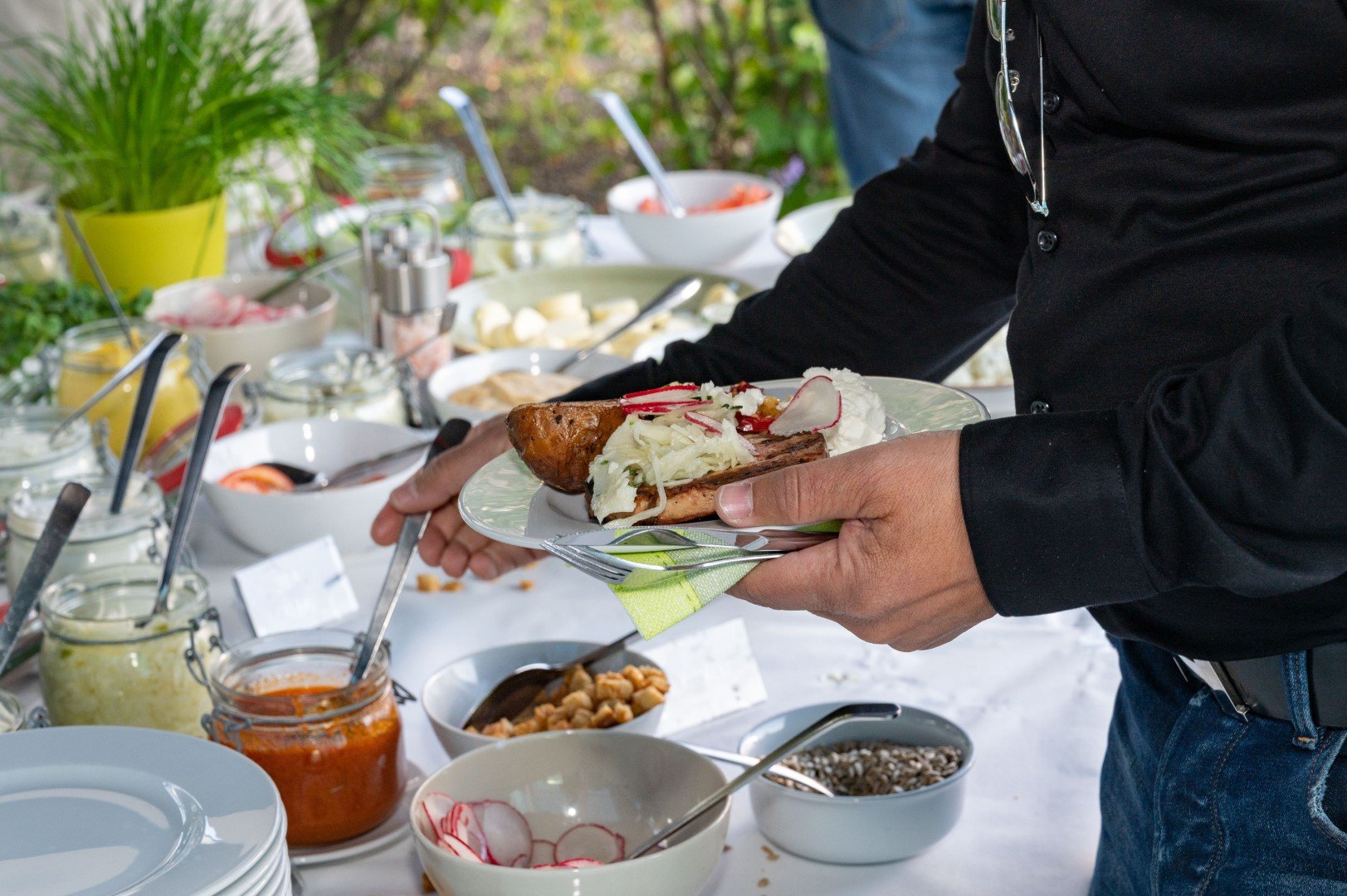 Person hält Teller mit Grillwurst, Sauerkraut und Radieschen bei einem Buffet.