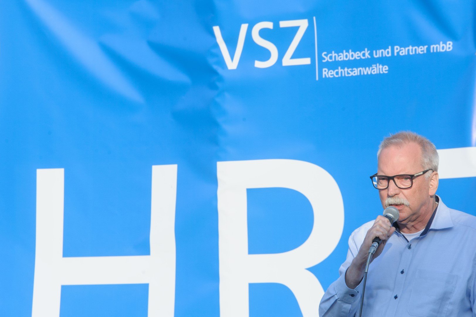 Ein älterer Mann mit Brille und grauem Haar spricht in ein Mikrofon vor einem blauen Hintergrund mit weißen Texten.
