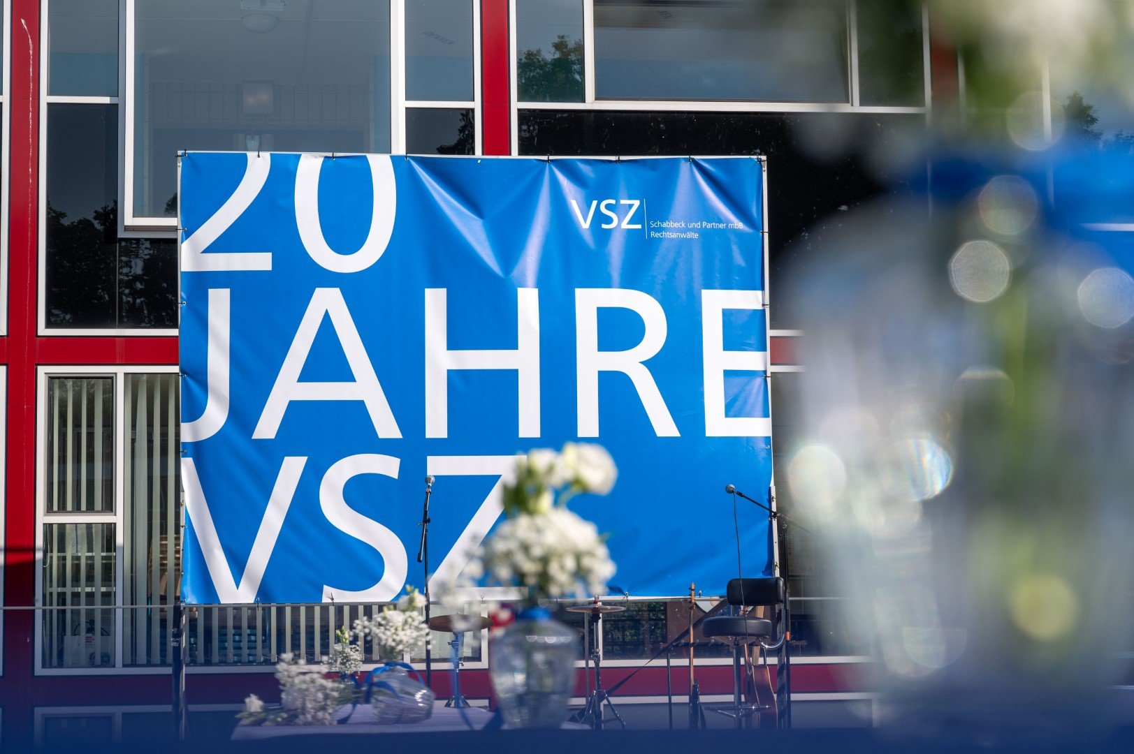 Blaue Jubiläumsfahne mit weißen Buchstaben, die '20 Jahre VSZ' lesen, vor einem modernen Gebäude mit roten und weißen Fensterrahmen.