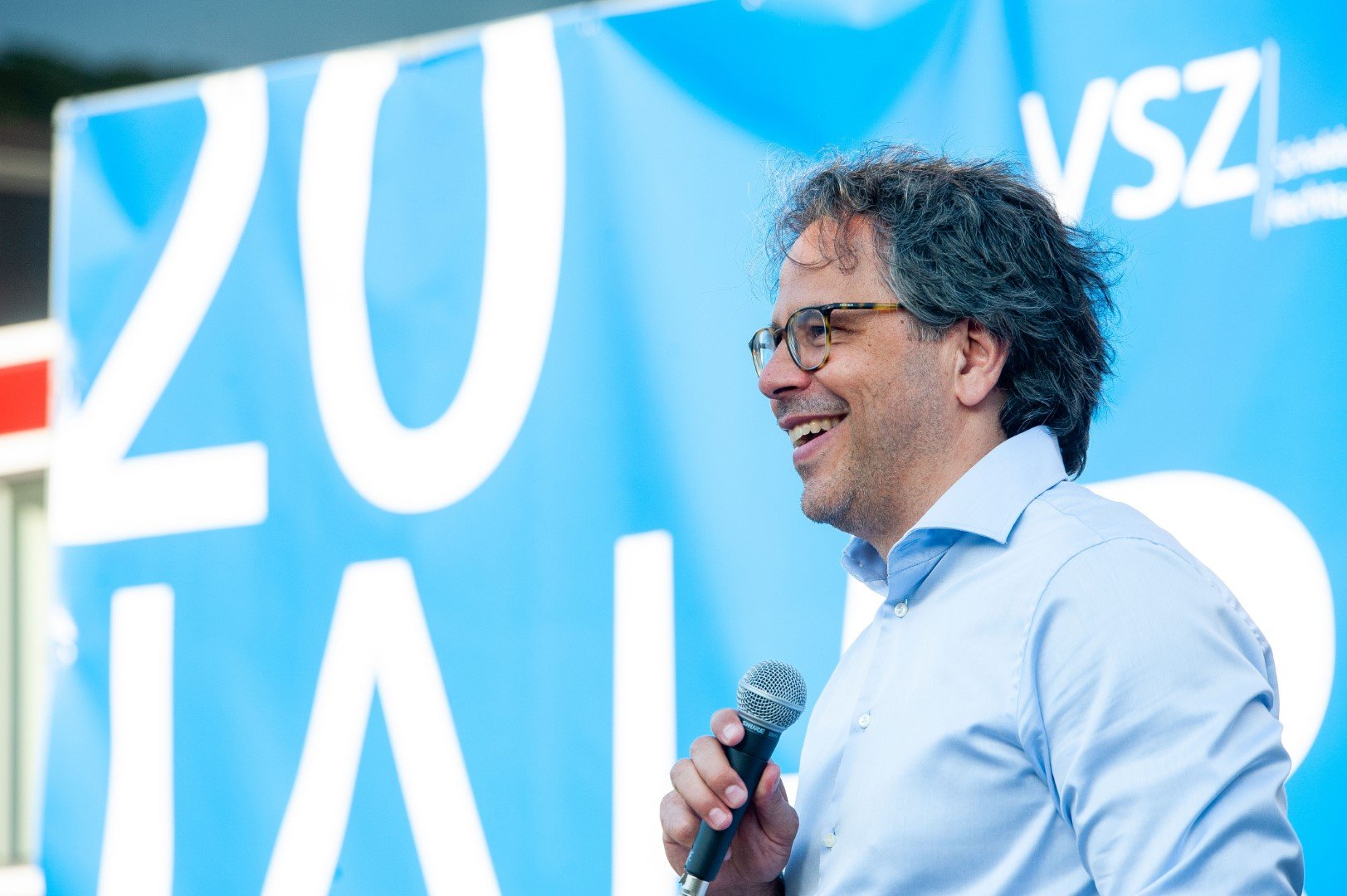 Jan Schabbeck von VSZ Rechtsanwälte spricht bei einer Veranstaltung, hält ein Mikrofon in der Hand, vor einem blauen Hintergrund mit weißen Buchstaben.