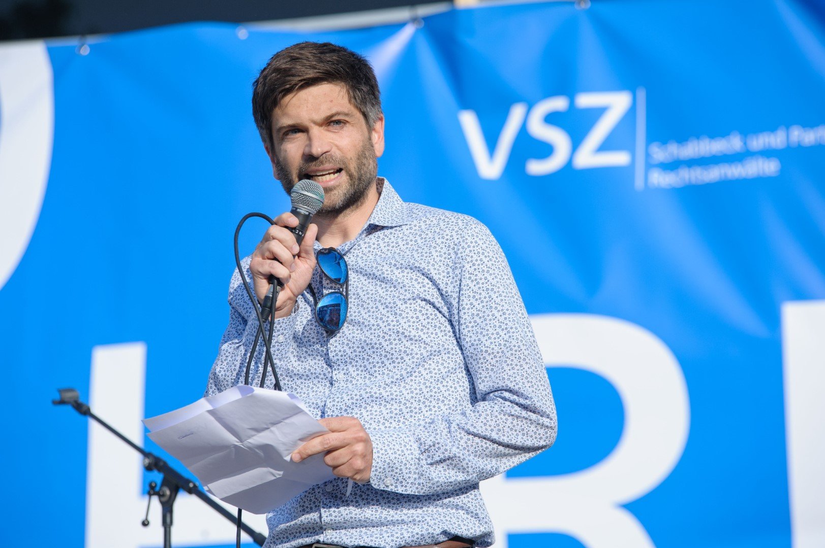 Karsten Mühlsteph von VSZ Rechtsanwälte spricht mit einem Mikrofon vor einer blauen Bühne, auf der das Logo der Partei 'VSZ' und die Worte 'Stadtstaat und Partei' zu sehen sind.