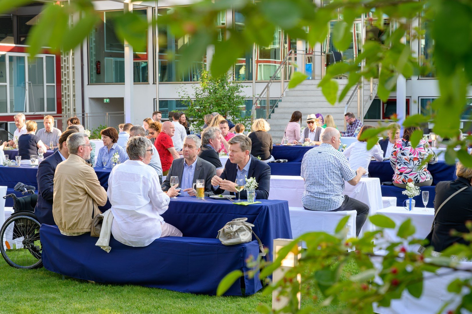 Menschen bei einem Outdoor-Gourmet-Essen, im Vordergrund durch Blätter beschattet, große Gebäude im Hintergrund.