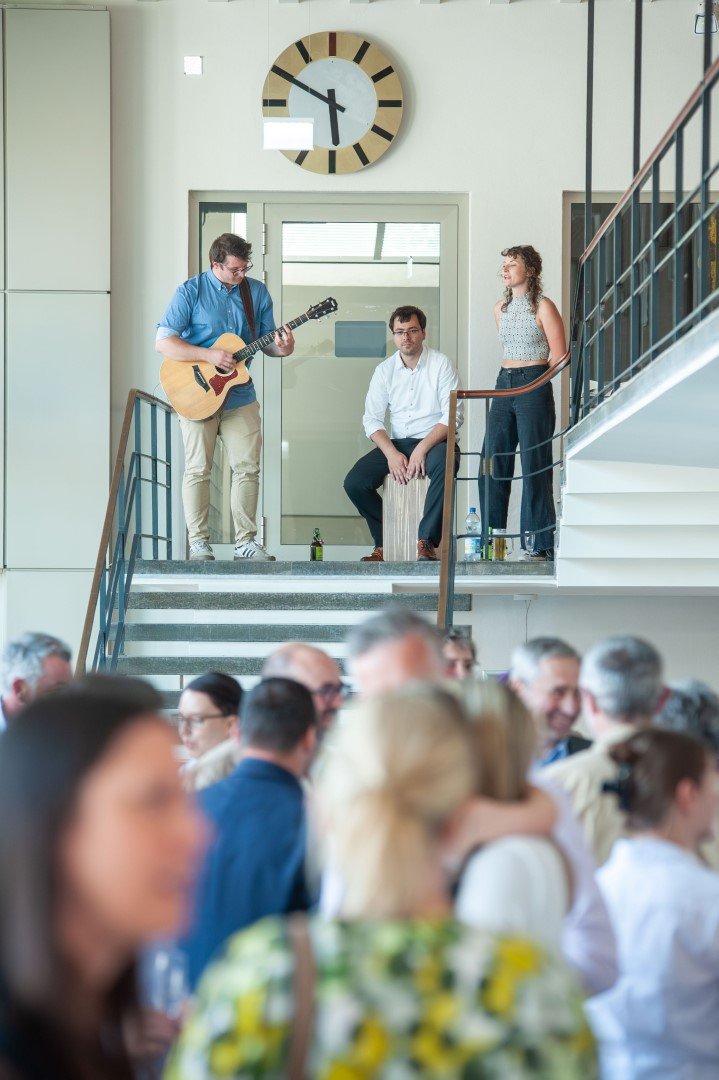 Drei Personen auf einer Treppe, einer spielt Gitarre, einer sitzt und die dritte steht, während im Hintergrund ein Publikum sitzt.