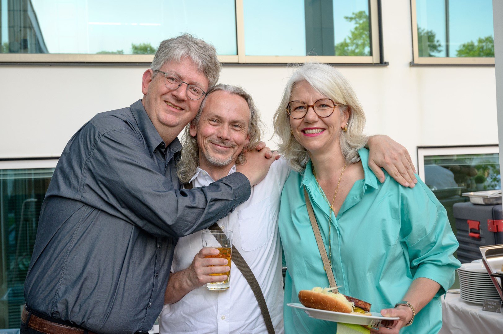 Drei lachende Erwachsene, zwei Männer und eine Frau, die sich umarmen und bei einer Feier im Freien sind. Die Frau hält einen Teller mit Hot Dogs, einer trinkt ein Bier.