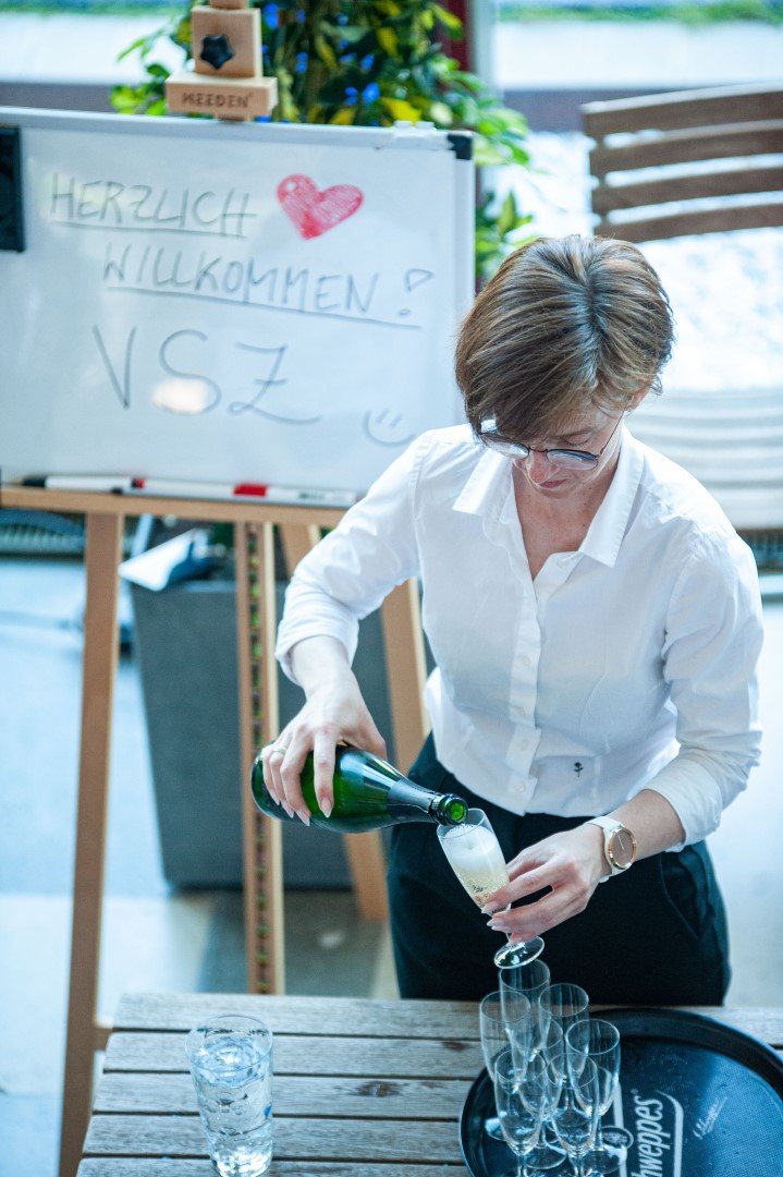 Eine Frau gießt Champagner in Gläser bei einer Party. Im Hintergrund eine Tafel mit der Aufschrift 'Herzlich Willkommen! V. S. Z'.