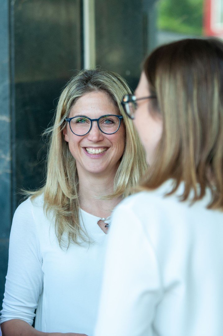 Zwei Frauen im Gespräch, eine lächelnd, die andere mit Brille, beide in Weiß, bei einem Treffen oder Gespräch im Freien.