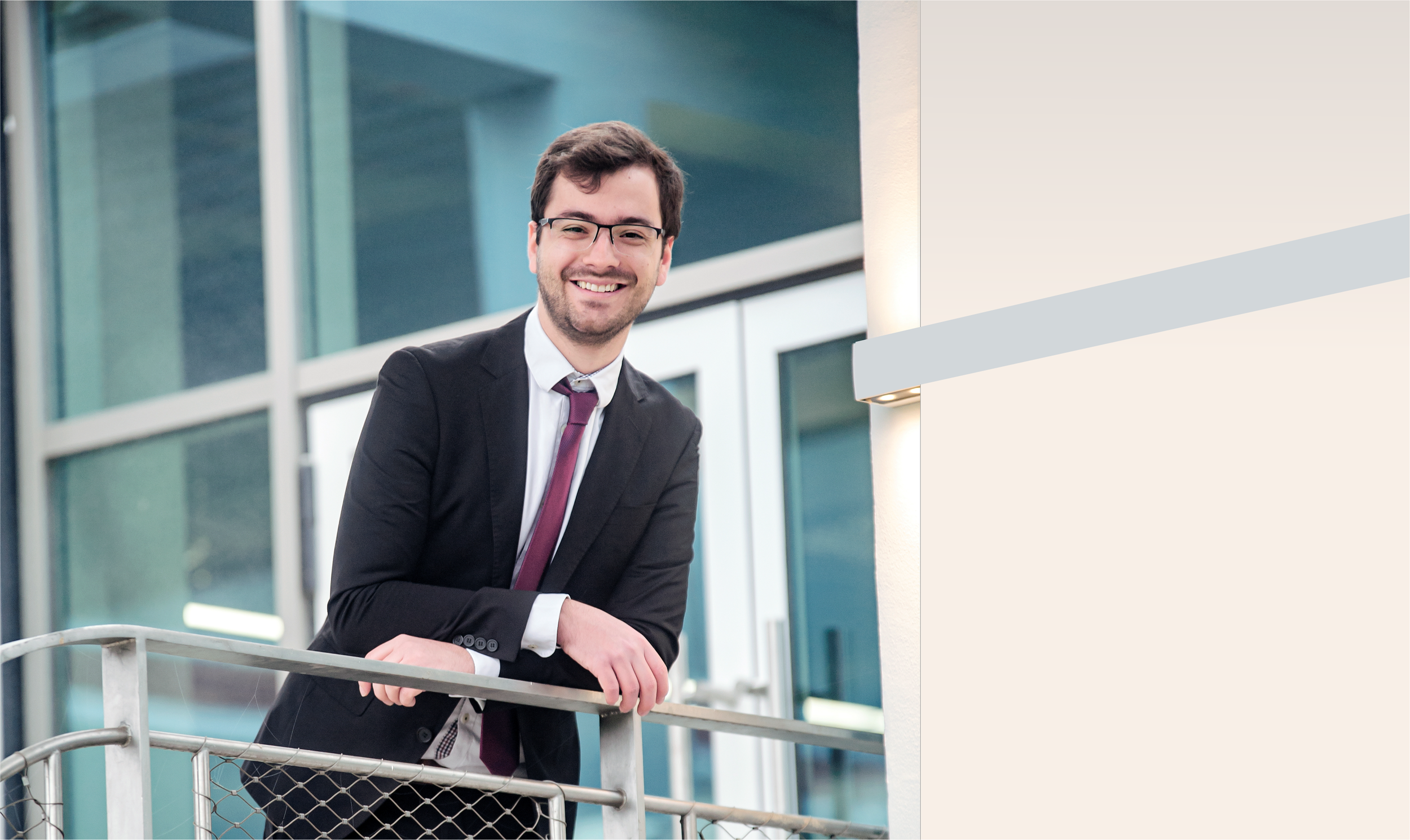 Ein lachender junger Mann in Geschäftskleidung steht vor einem großen Bürogebäude, der Blick nach vorne gerichtet.