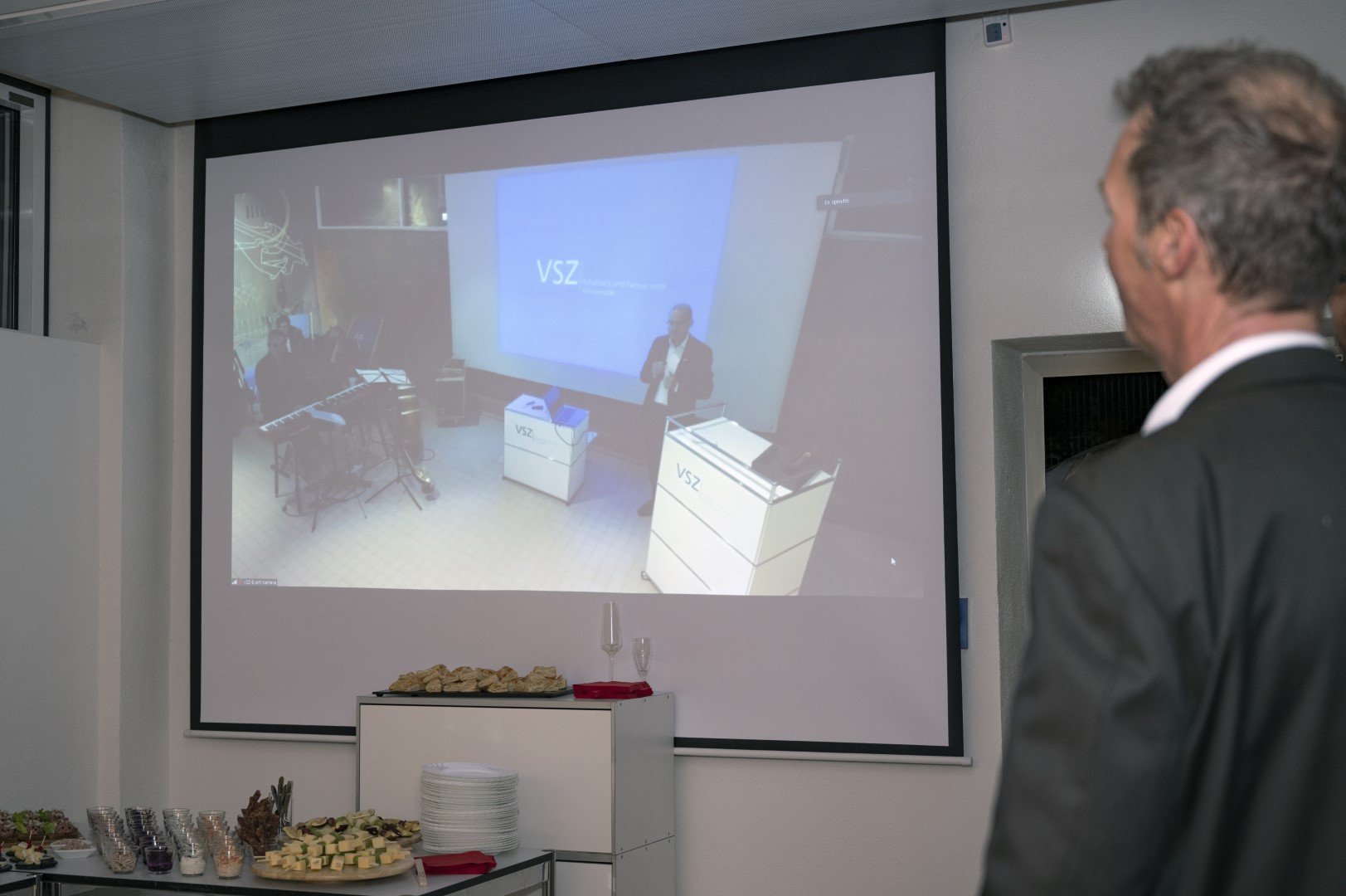 Blick auf eine Präsentation im Saal mit Mensch im Anzug und auf der Leinwand. Buffet mit Getränken und Snacks im Vordergrund.