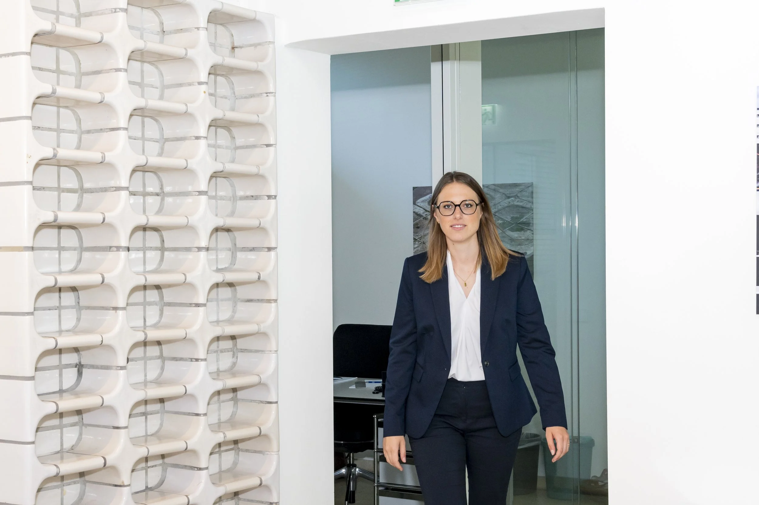 Eine Frau in dunklem Anzug und weißem Hemd steht in einem modernen Büro, im Hintergrund eine Glastür und eine Wand mit weißem, perforiertem Design.