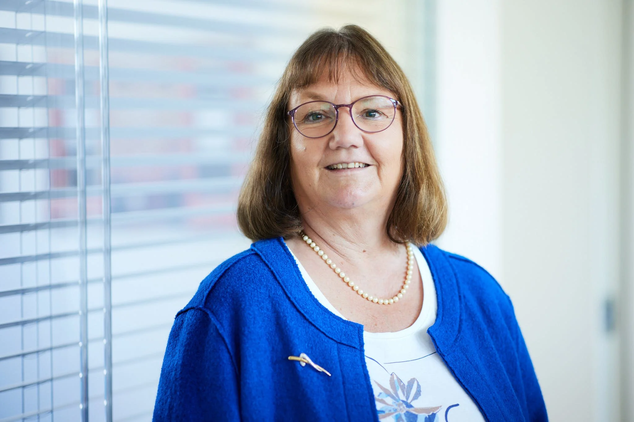 RAin Zickgraf von VSZ Rechtsanwälte trägt ein blaues Jacke und eine Perlenkette, im Hintergrund eine Fensterjalousie.