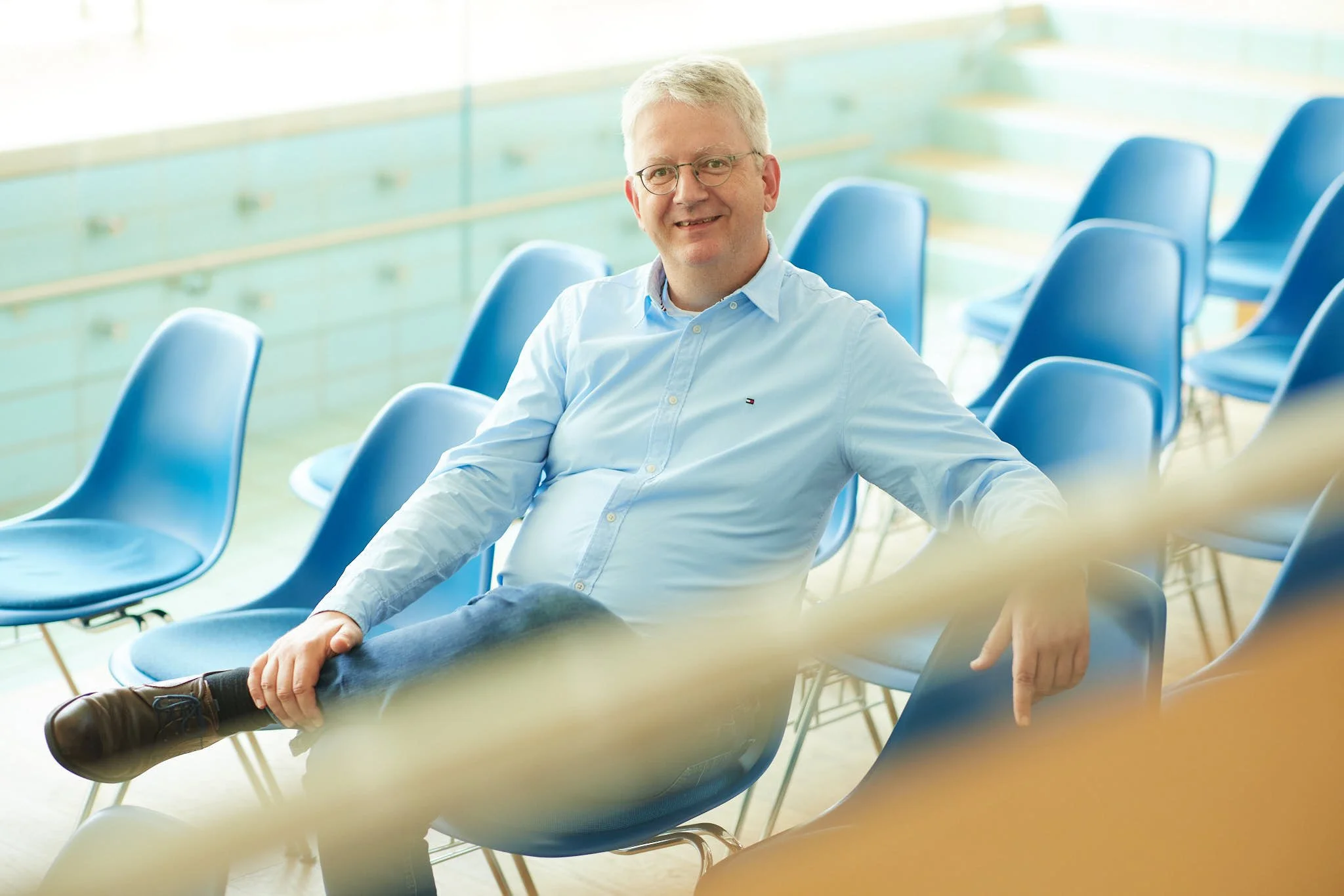 Daniel Hamers von VSZ Rechtsanwälte sitzt in einem Raum mit blauen Stühlen, vermutlich in einem Auditorium oder Wartebereich, der im Hintergrund gefilmt wurde.