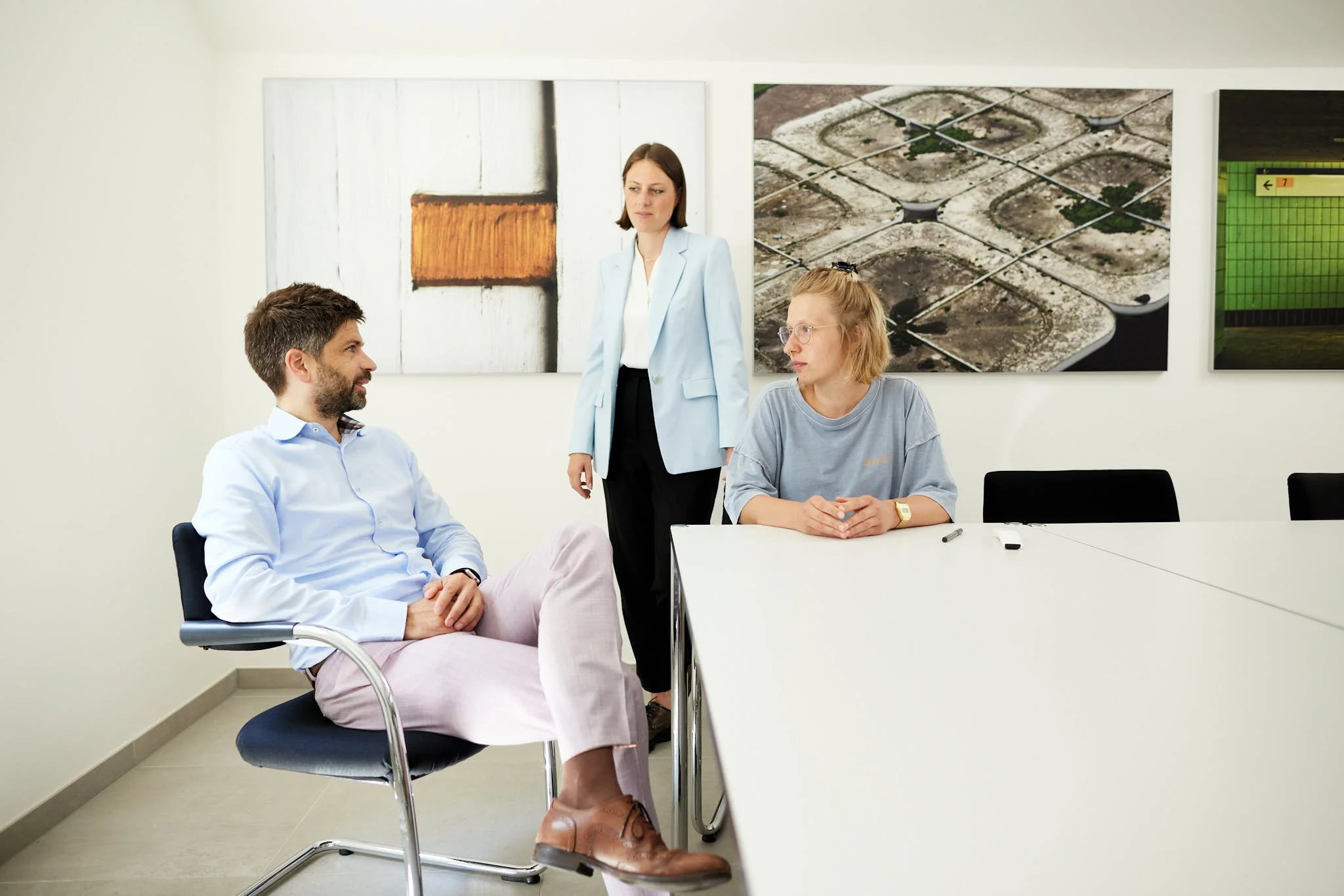 VSZ Rechtsanwälte in einem Konferenzraum, zwei sitzen am Tisch, eine steht. Im Hintergrund sind drei Wandbilder mit Kunstwerken.