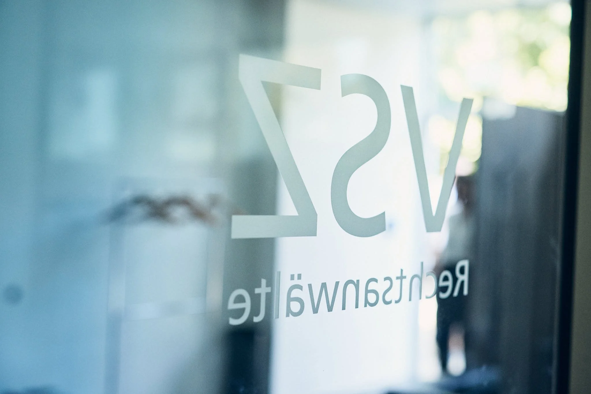 Auf einer Glasoberfläche sind weiße Buchstaben zu sehen, die teilweise durch das Glas spiegeln. Der Hintergrund ist unscharf mit natürlichem Licht, das durch Fenster scheint, und es sind verschwommene grüne und helle Bereiche erkennbar.