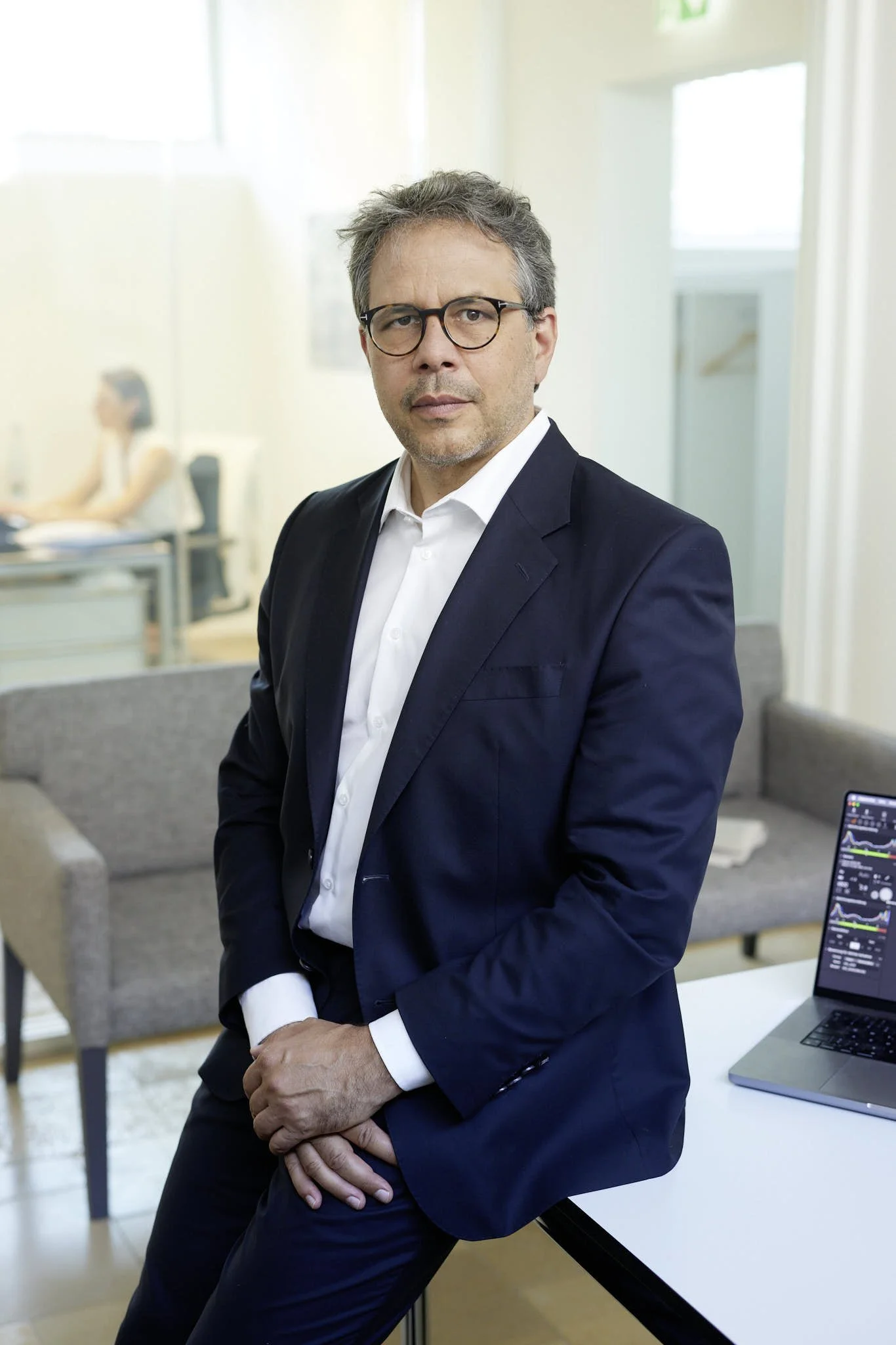 RA Jan Schabbeck von VSZ Rechtsanwälte in einem dunklen Anzug und weißen Hemd, mit Brille, steht in einem modernen Büro. Im Hintergrund sind ein Sofa, ein Tisch, ein Laptop mit Diagrammen und eine Person, die mit einem Handy arbeitet.