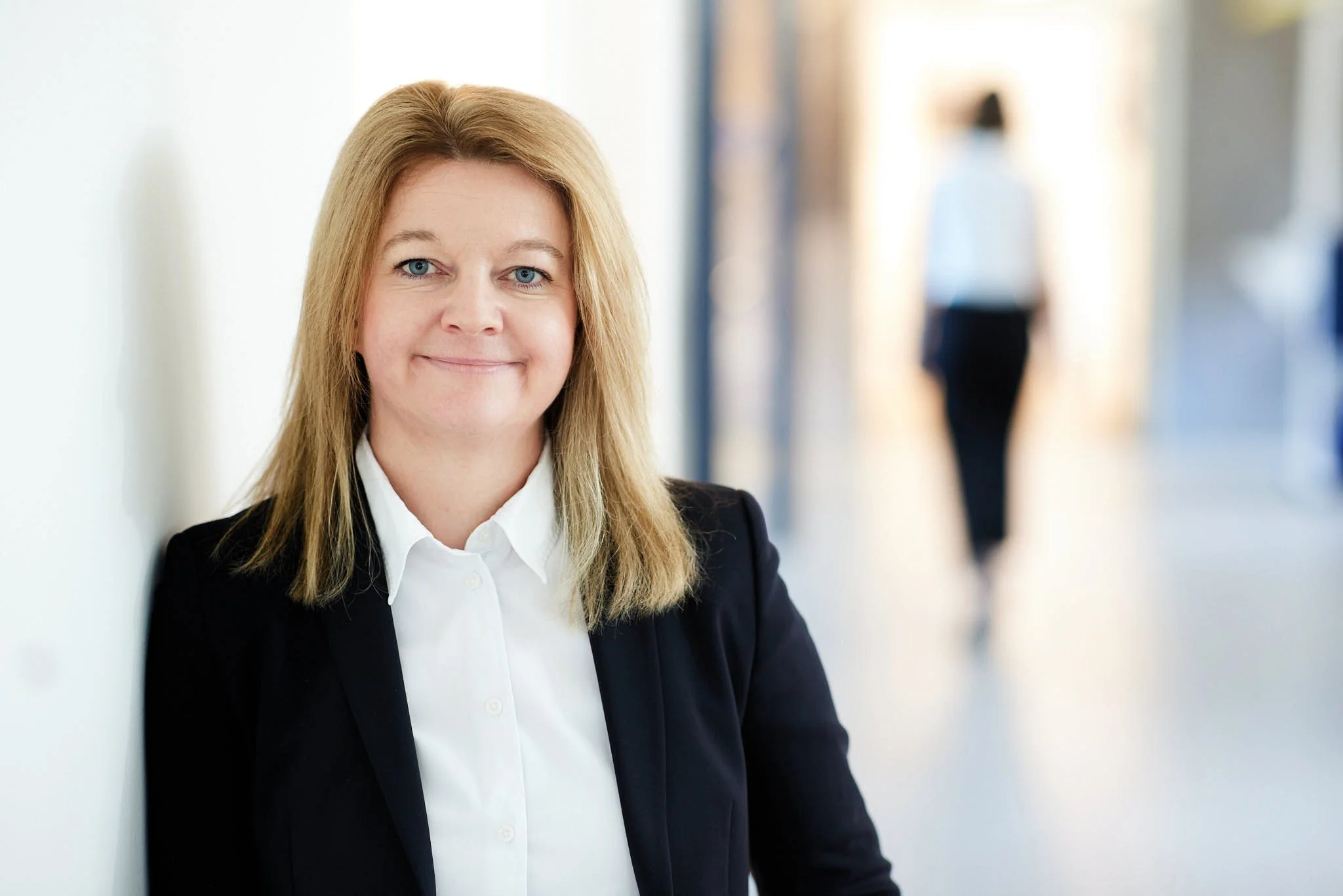 RAin Angela König von VSZ Rechtsanwälte lächelt, während sie an einer weißen Wand lehnt, im Hintergrund sind verschwommene Figuren in einem modernen Büro.