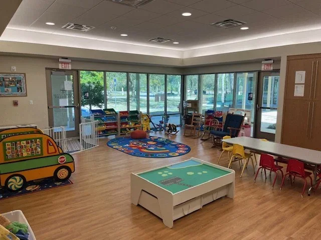 A spacious preschool or daycare room with large windows, colorful toys, a patterned rug, small tables and chairs, and a play area.