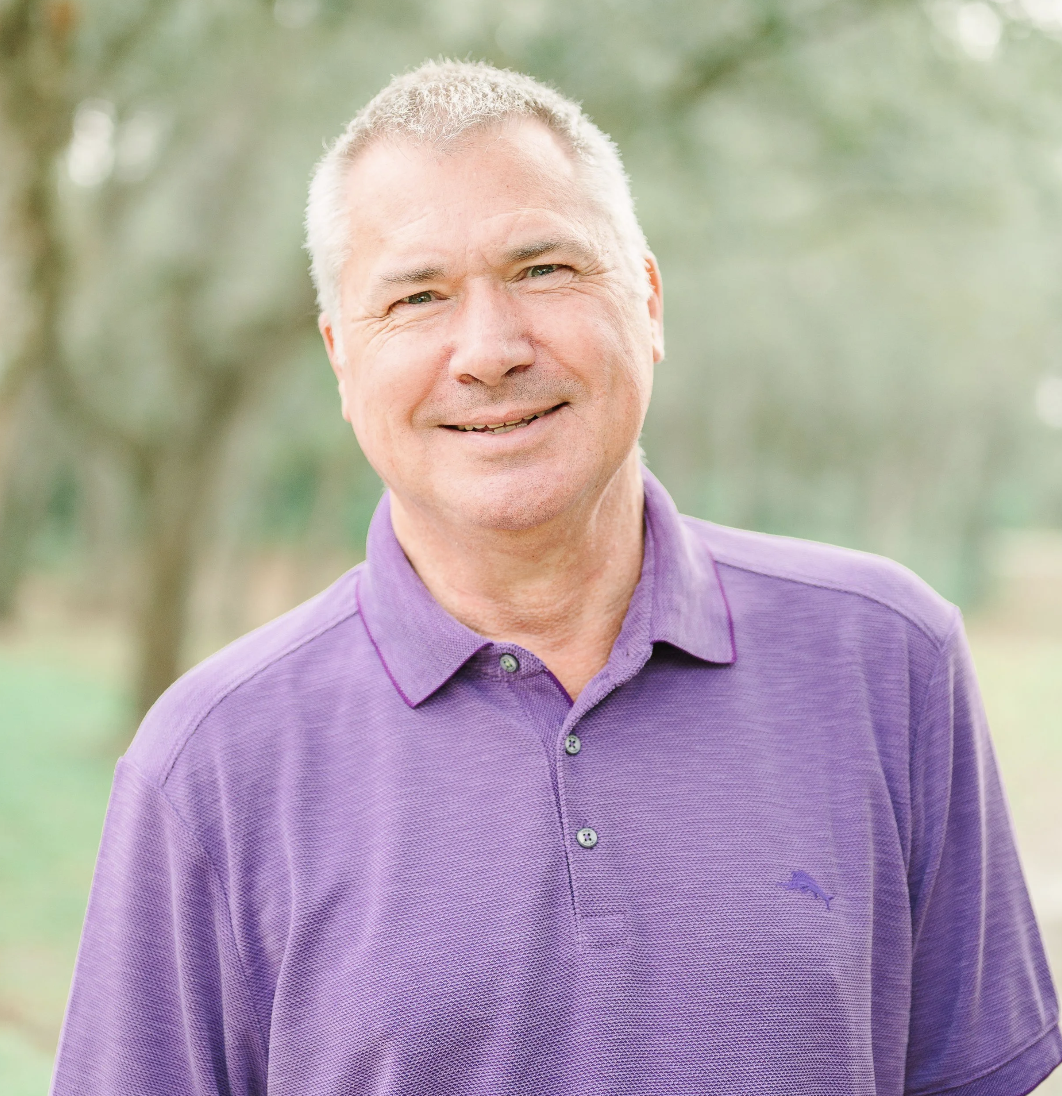 A middle-aged man with gray hair smiling outdoors, wearing a purple polo shirt with a small logo on the chest, in a park-like setting with trees in the background.