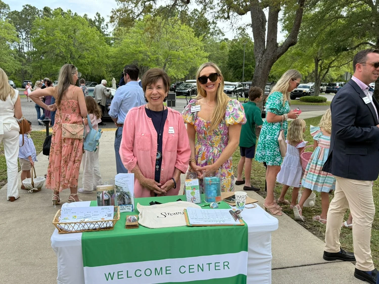 Two women standing behind a table with a 'Welcome Center' sign at an outdoor event, surrounded by people, trees, and parked cars.