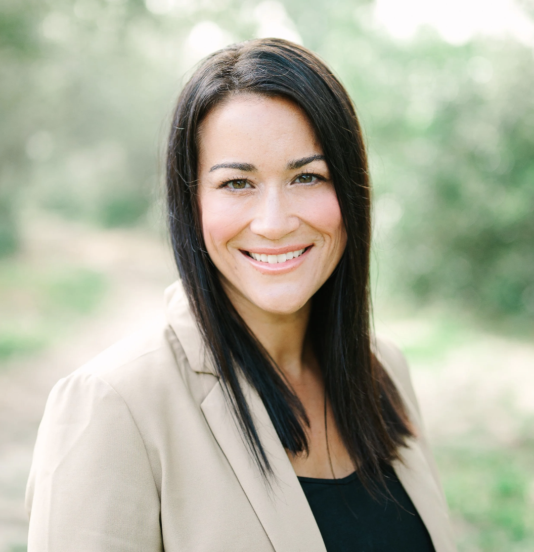 Portrait of a woman smiling outdoors with blurred greenery in the background.
