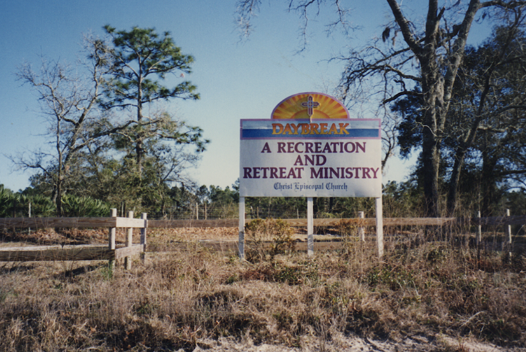 Sign with sunrise and cross, advertising daybreak recreation and retreat ministry at Christ Episcopal Church, set in a rural outdoor area with trees and fence.
