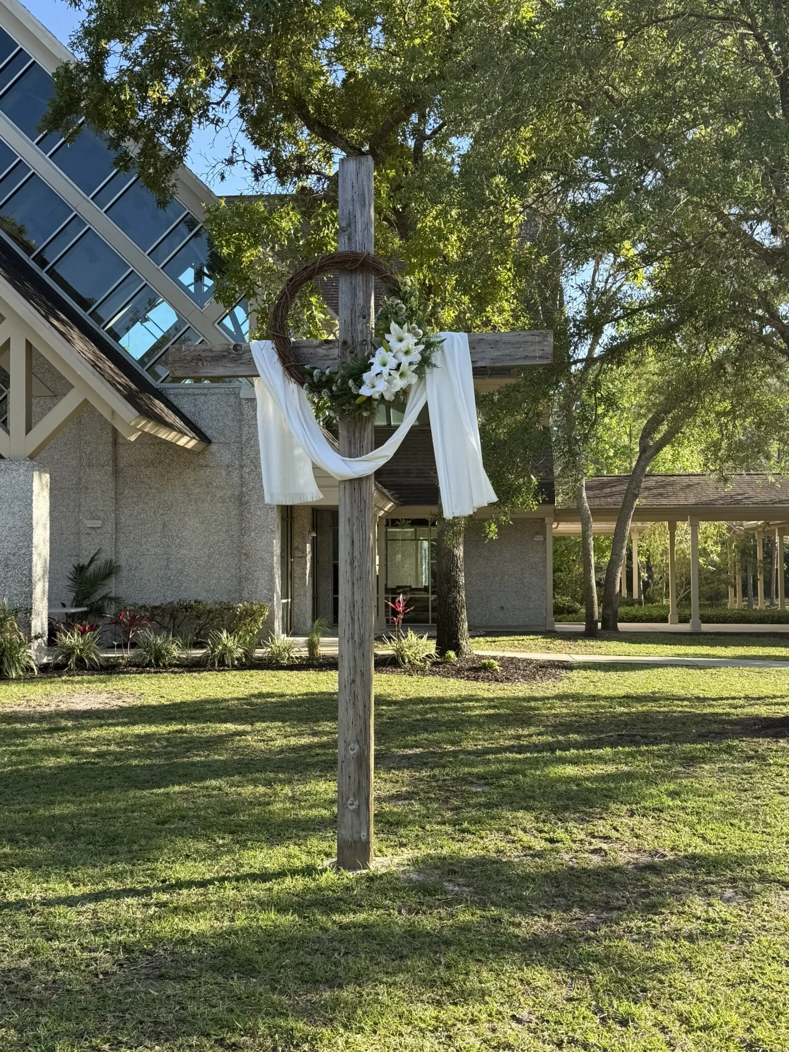 A wooden cross decorated with white cloth, white lilies, and greenery, in an outdoor setting with trees and a building in the background.