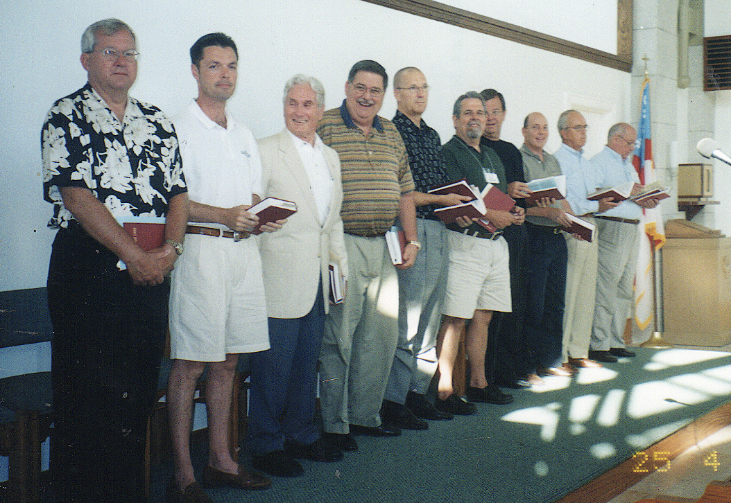 A group of ten men standing on a stage in front of a white wall, holding books and dressed in casual and semi-formal attire, during a ceremony or presentation.