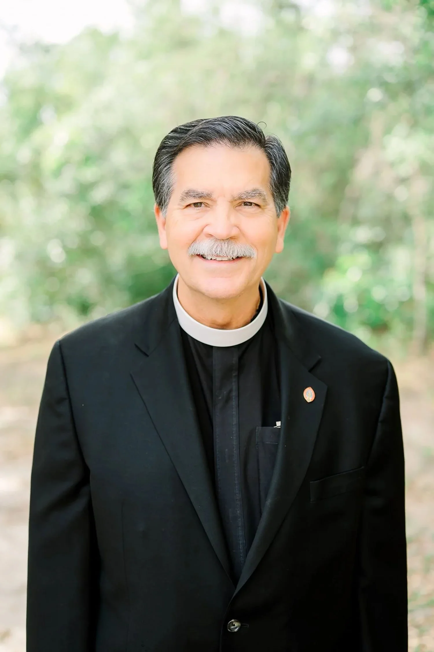 A smiling middle-aged man with dark hair and a white mustache, wearing black clerical attire with a white collar, standing outdoors with a blurred green background.