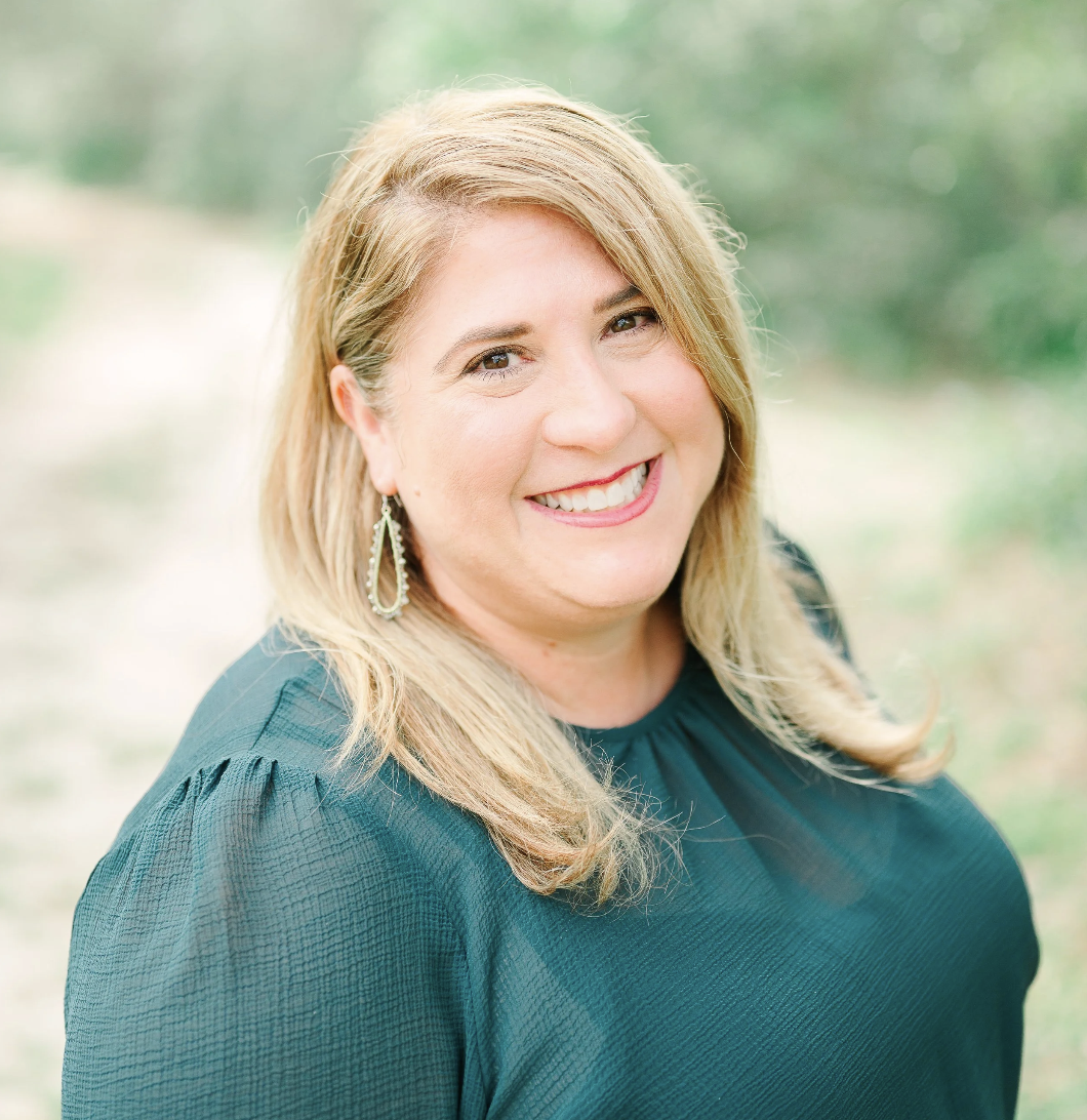 Portrait of a smiling woman with blonde hair, wearing a black textured top and silver earrings, outdoors with blurred nature background.