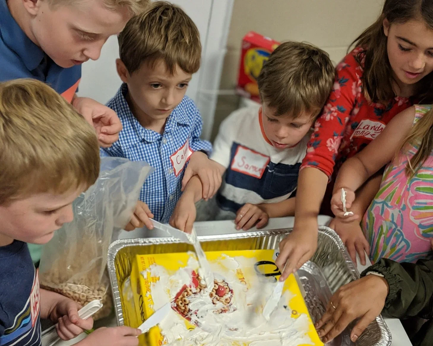 Children gathered around a table cutting into a birthday cake, some with whipped cream and strawberries, in a classroom or party setting, with stickers on their shirts.