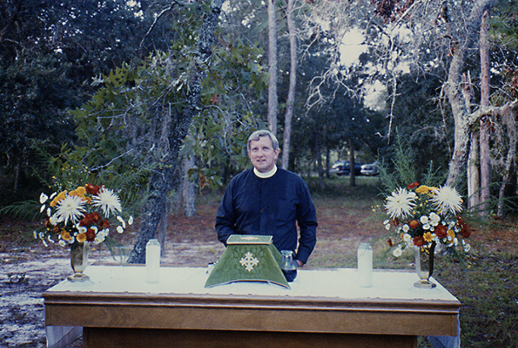 A man in a black jacket stands behind an outdoor altar with two flower arrangements, candles, and a green cloth with a gold cross, set in a wooded area.