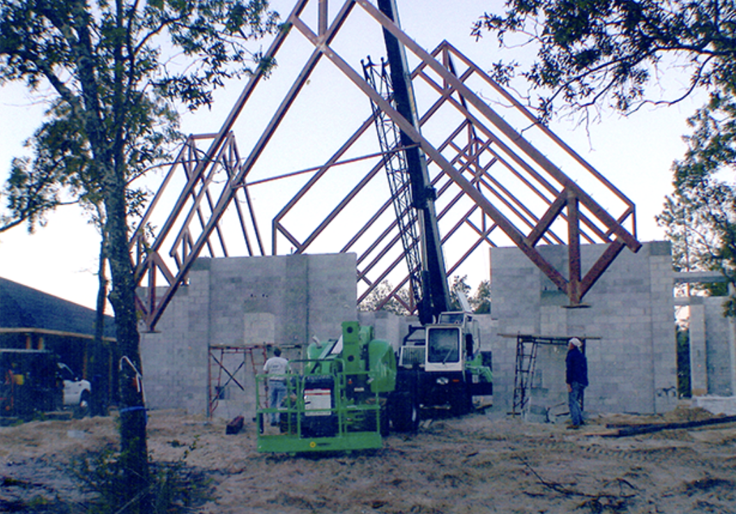 Construction site with a crane lifting a large red steel roof frame over gray stone walls, two workers supervising, and trees surrounding the site.