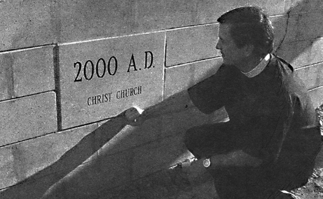 A woman and a man are kneeling next to a memorial wall inscribed with '2000 A.D.', 'CHRIST CHURCH'. The woman is helping the man plant or place a flower or object into the ground.