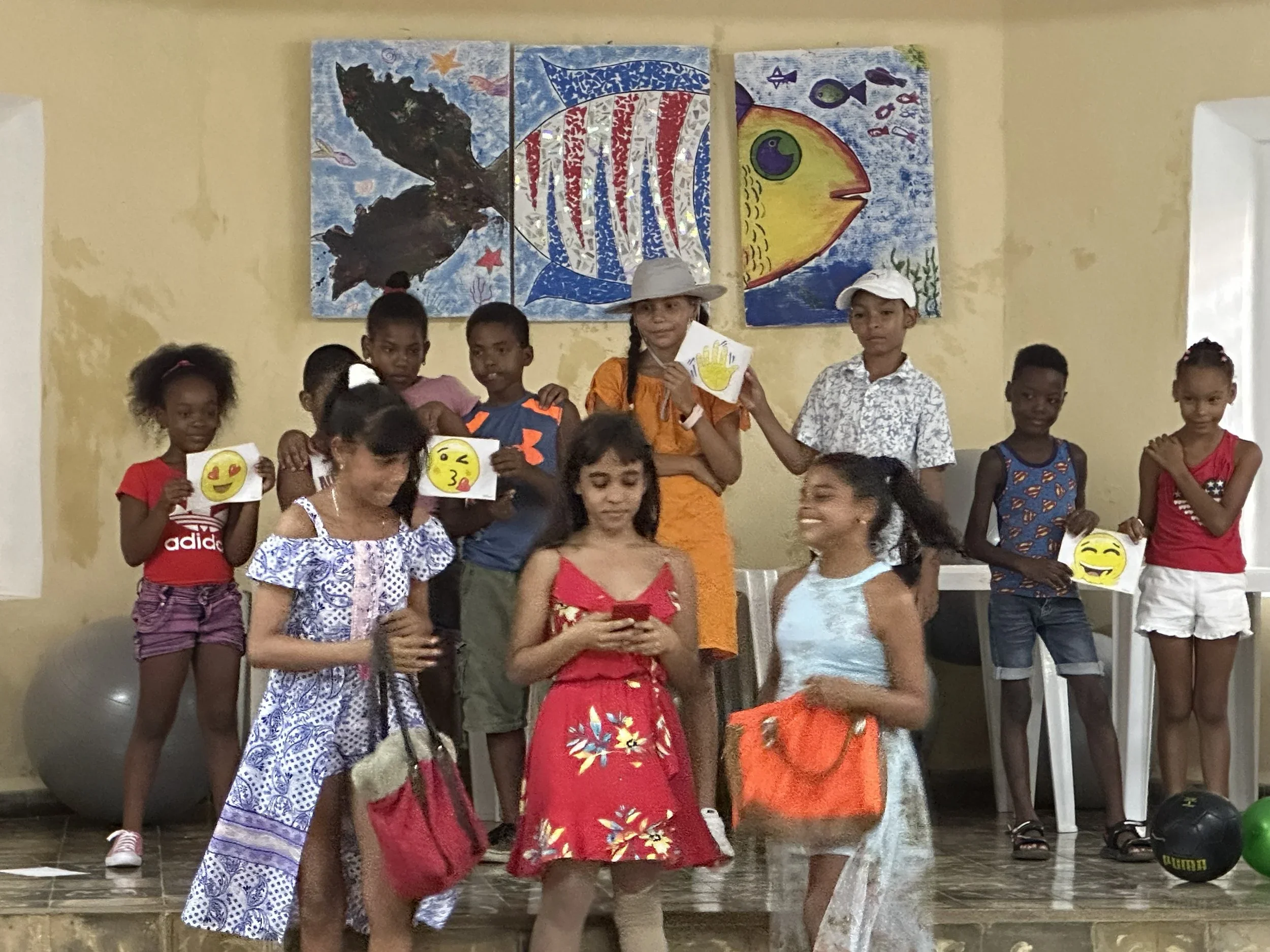 A group of young children, some holding emoji-themed paper fans, standing and sitting in what appears to be a classroom, with colorful underwater-themed art on the wall behind them.