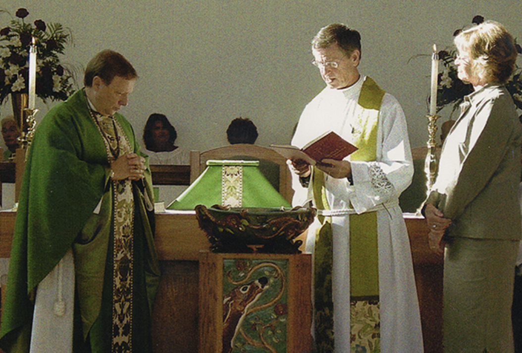 A religious ceremony with three priests, including a woman in green robes, in front of a altar with candles, flowers, and religious symbols.