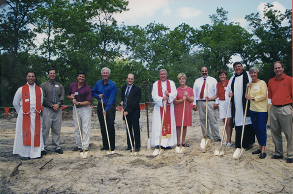 Group of people at a groundbreaking ceremony holding shovels on a dirt site, with trees in the background.