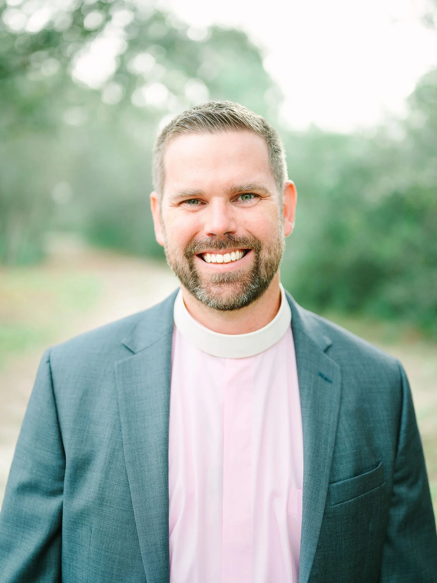 A man smiling outdoors wearing a suit and white clerical collar.