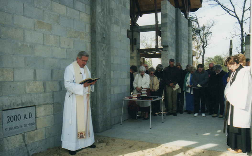 A priest leading a congregation prayer outside a church under construction, with a sign reading '2000 A.D. St. Francis In-The-Field'.
