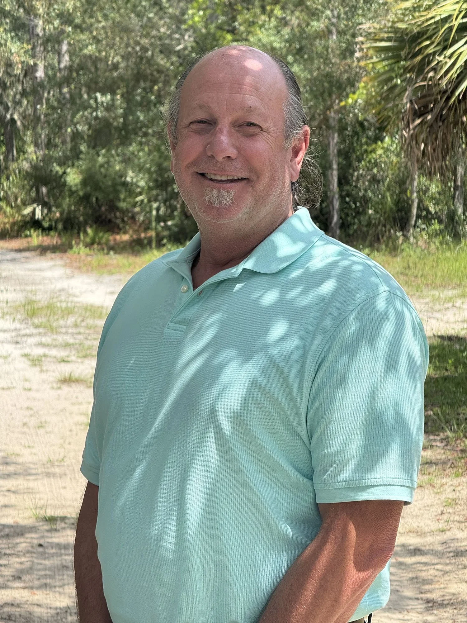 A smiling man with a bald head and a goatee, standing outdoors in a setting with trees and greenery, wearing a light blue collared shirt.