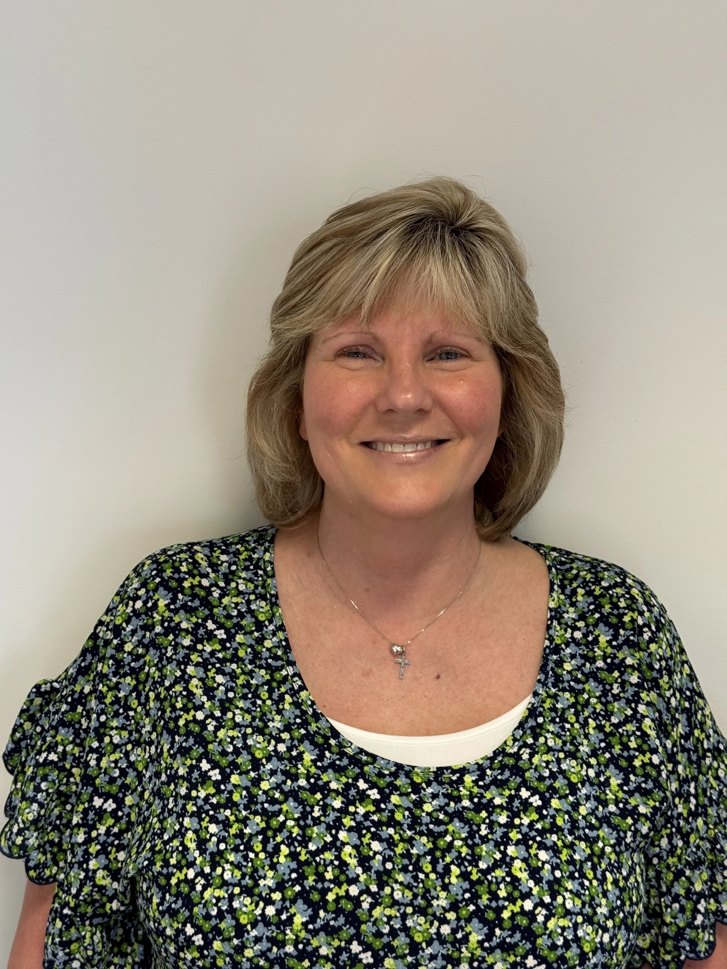 A smiling woman with shoulder-length blonde hair, wearing a black dress with a small white and green floral pattern, a white undershirt, and a silver necklace with a cross pendant, standing against a plain white wall.