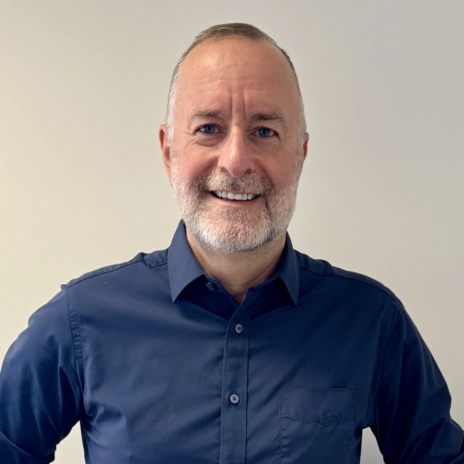 A smiling middle-aged man with gray hair and beard wearing a white button-down shirt against a plain light-colored wall.