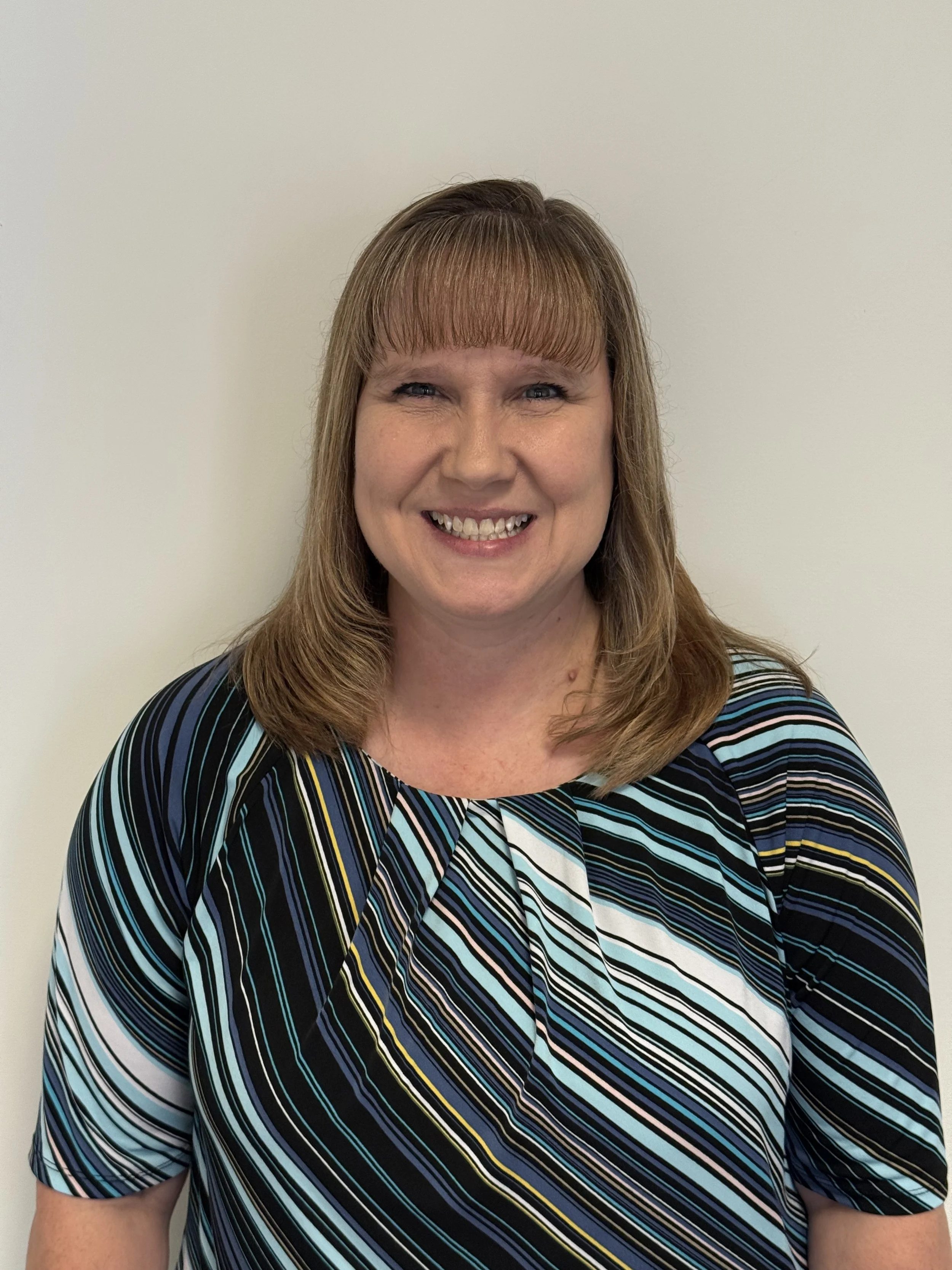 A woman with shoulder-length light brown hair, bangs, and a big smile, wearing a colorful striped top standing against a plain white wall.