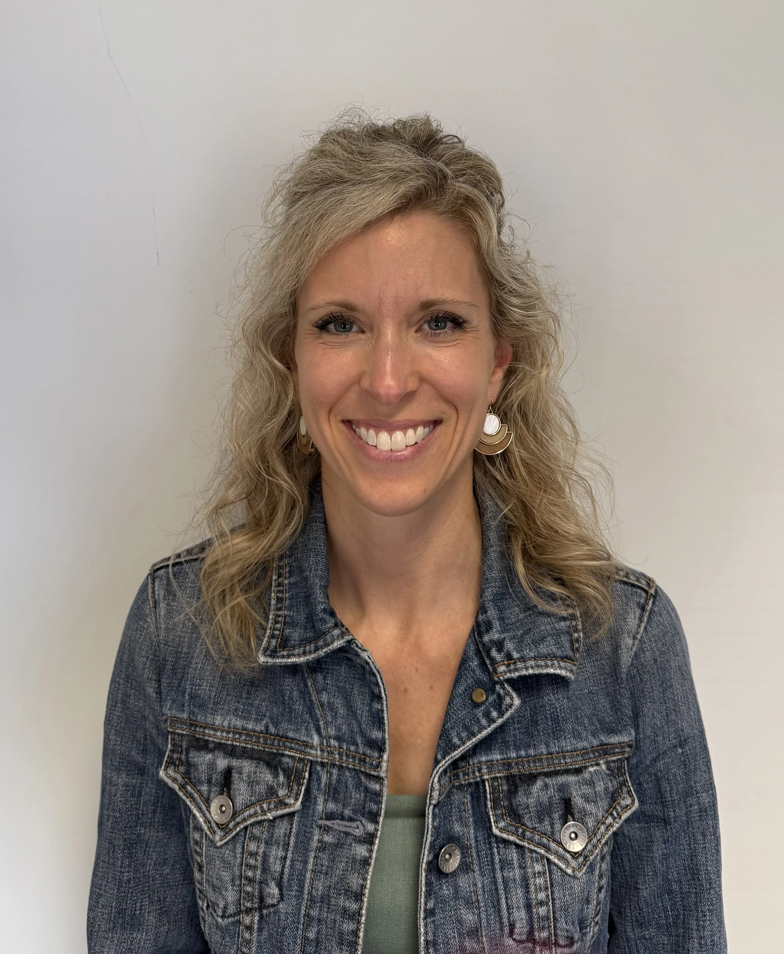 A woman with curly blonde hair smiling at the camera, wearing a denim jacket and earrings, standing against a plain white wall.