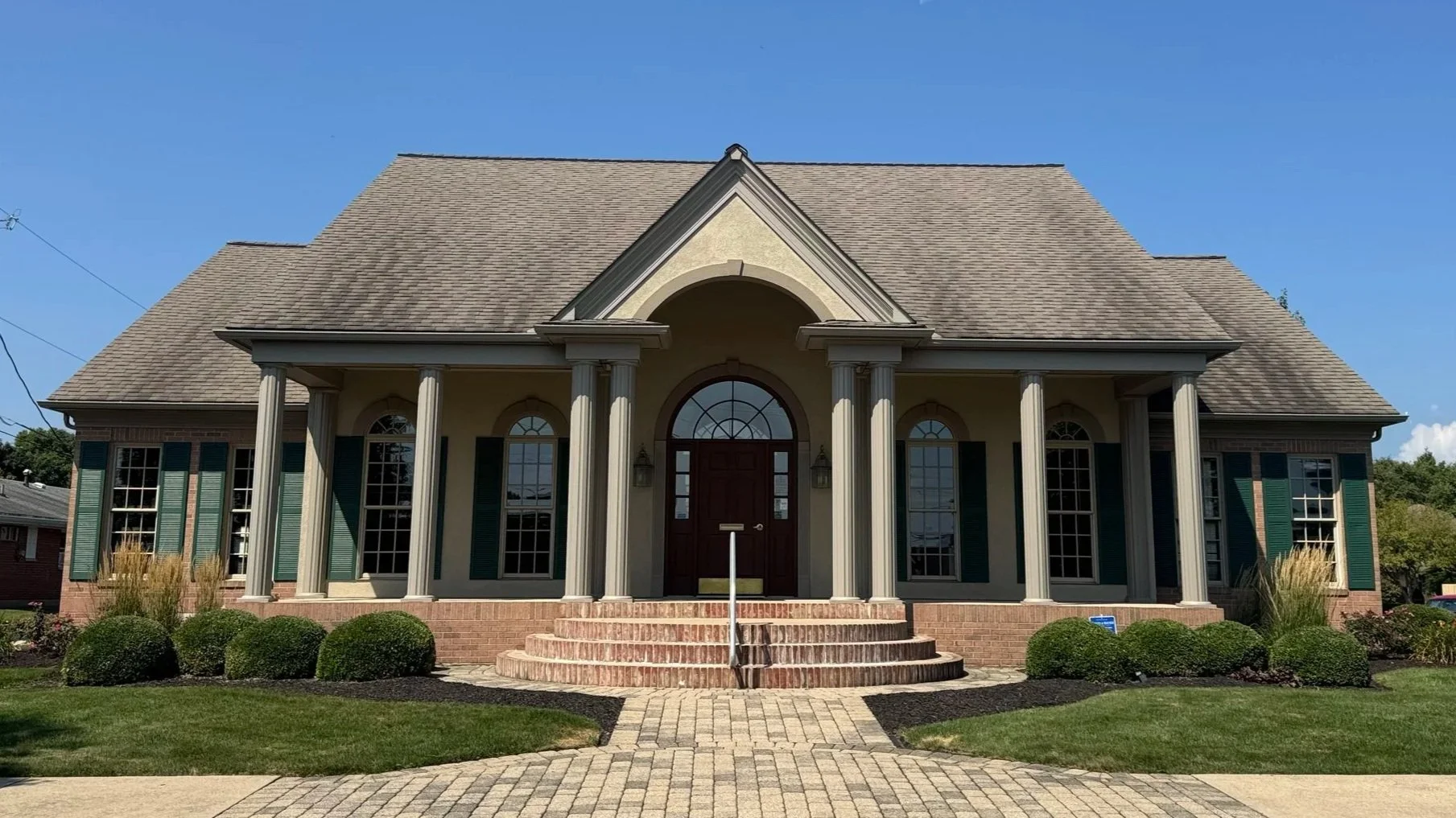 A large, two-story house with a symmetrical front, brick stairs, and columns supporting a portico. The house has multiple tall windows with green shutters and a gable roof. The front yard features a paved walkway, trimmed bushes, and landscaped garden beds.