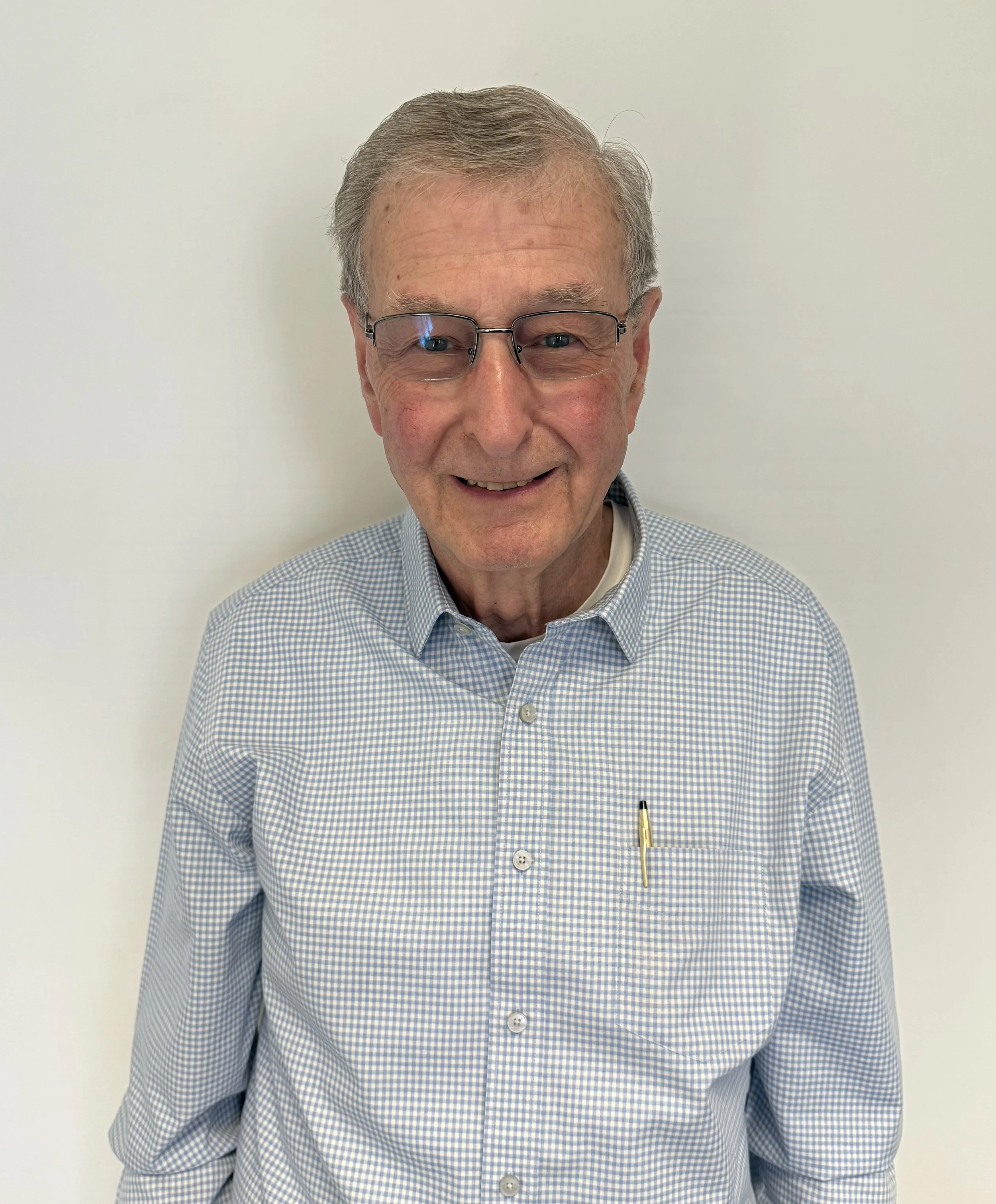 An elderly man with glasses smiling, wearing a blue and white checkered shirt, standing against a plain white background.