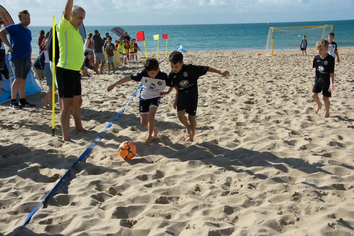 Kids playing beach soccer on sandy shore with ocean in background, spectators and other children nearby, clear sky.